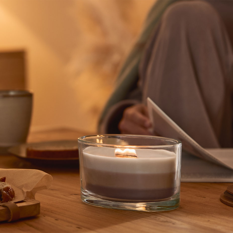 Brennende dreischichtige Kerze in einem Glas, mit einer lesenden Person im Hintergrund.