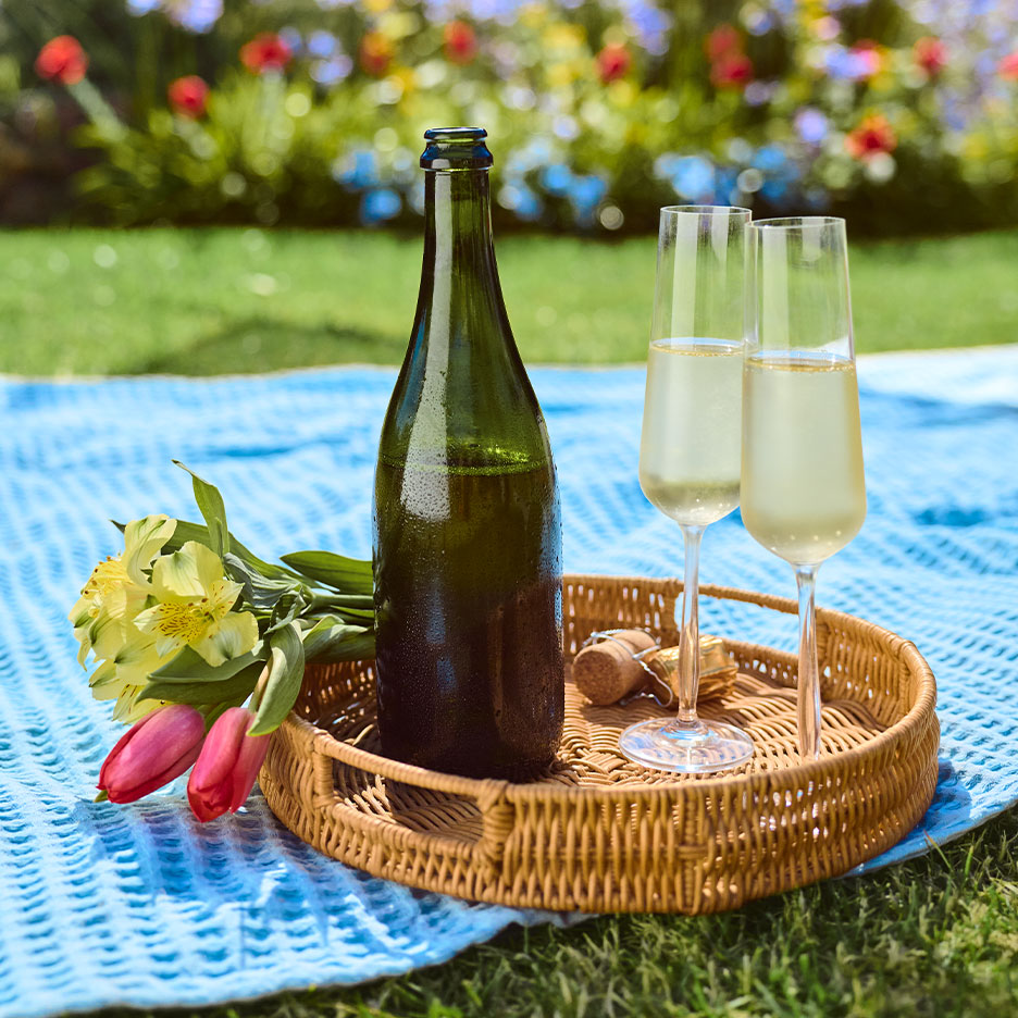 Sektflasche und zwei Gläser auf einem Tablett mit Blumen auf einer Picknickdecke im Gras.