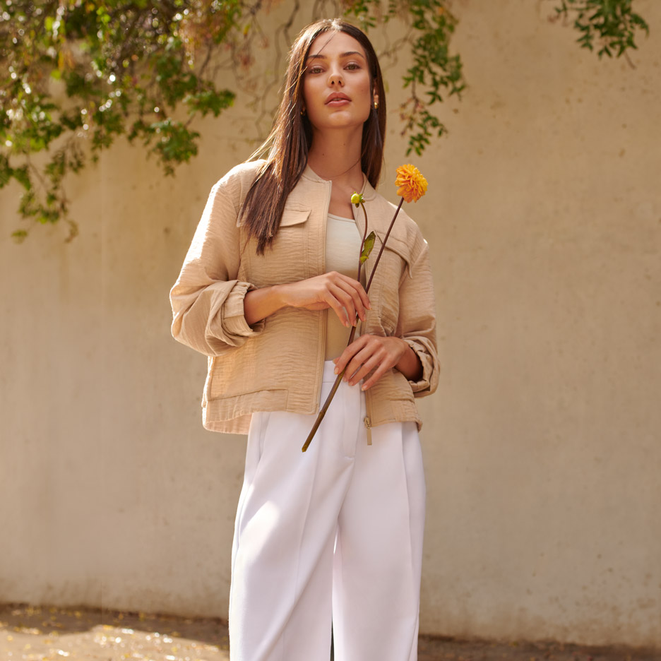Frau in beiger Jacke und weißer Hose, die eine orangefarbene Blume hält.