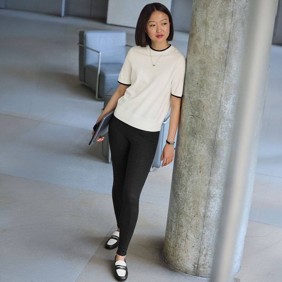 Frau in weißem Kurzarm-Top mit schwarzem Rand, schwarzen Leggings und zweifarbigen Loafern.