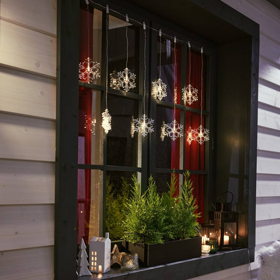 Schneeflocken-Lichterkette hängt in einem Fenster mit kleinen Tannen und Kerzen auf der Fensterbank.