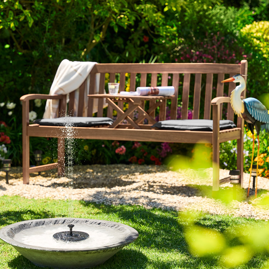 Gartenbank aus Holz mit Klapptisch, Kissen und Solarbrunnen in einem sonnigen Garten.