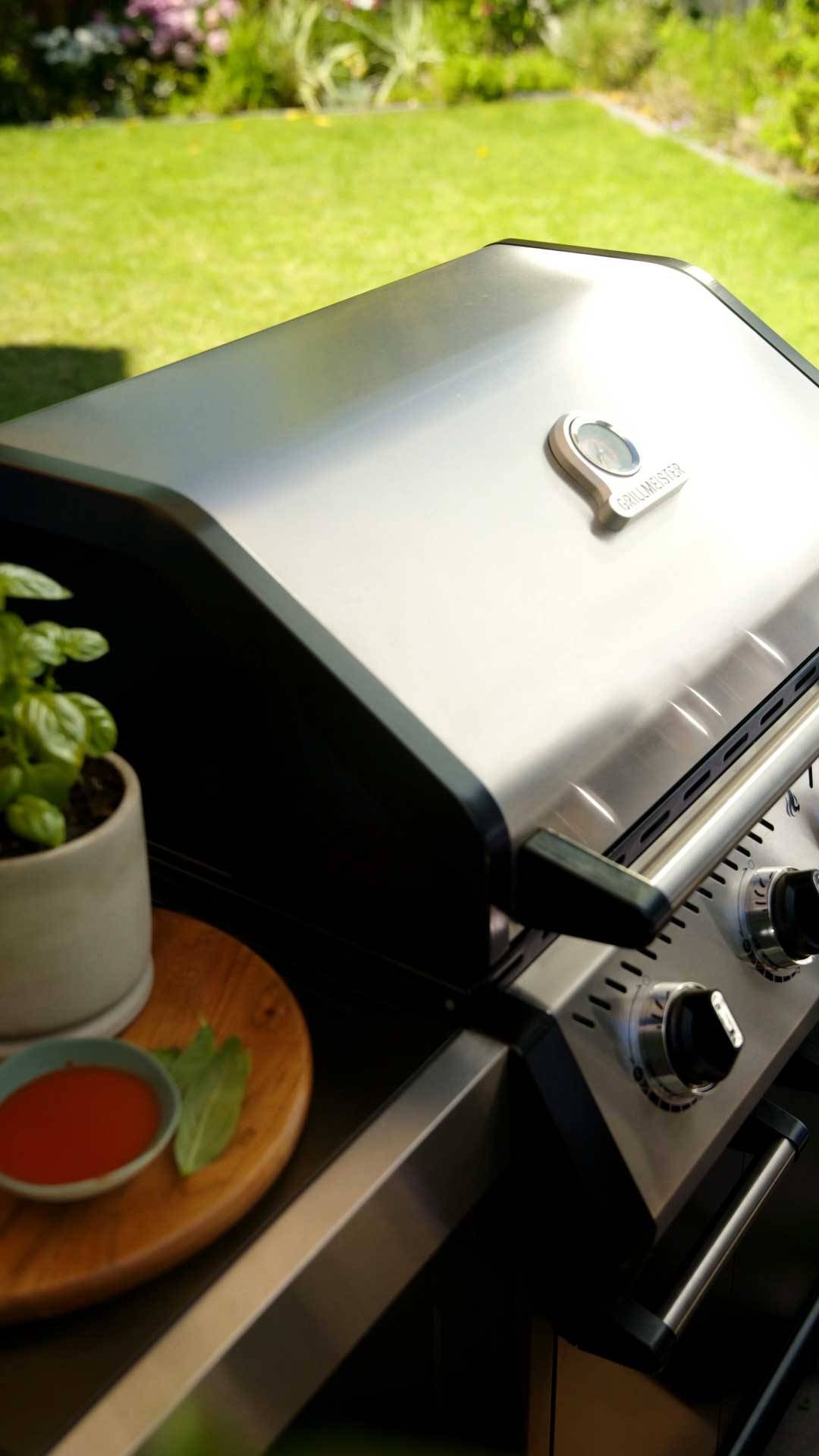 Gasgrill aus Edelstahl mit Thermometer, daneben ein Basilikumtopf und Soße.