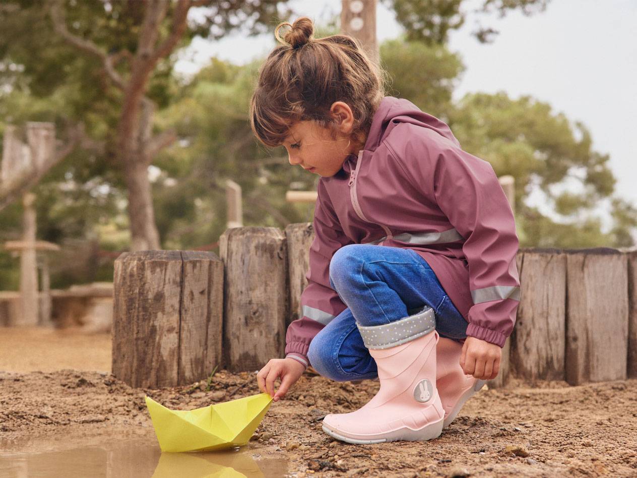 Mädchen in rosa Gummistiefeln und Jacke spielt mit einem Papierboot in einer Pfütze.