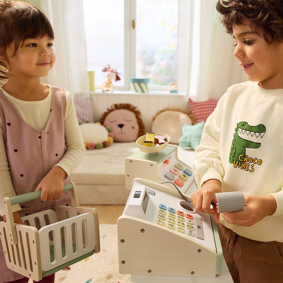 Kinder spielen Kaufladen; Junge bedient Registrierkasse, Mädchen trägt Einkaufskorb.