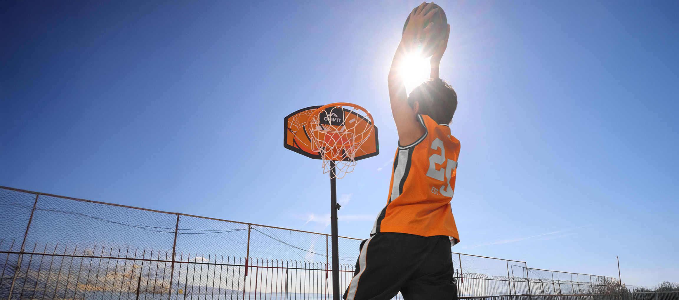Junge in orangefarbenem Trikot wirft Ball in Basketballkorb vor blauem Himmel.