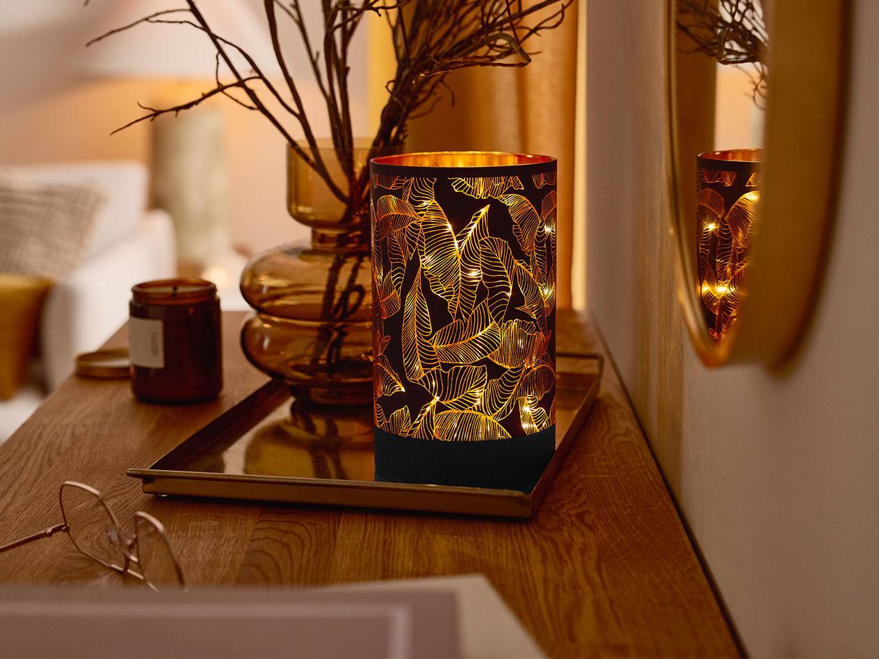 Eine dekorative LED-Laterne mit Blattmuster, eine Vase mit Ästen und eine Kerze auf einem Holztisch.