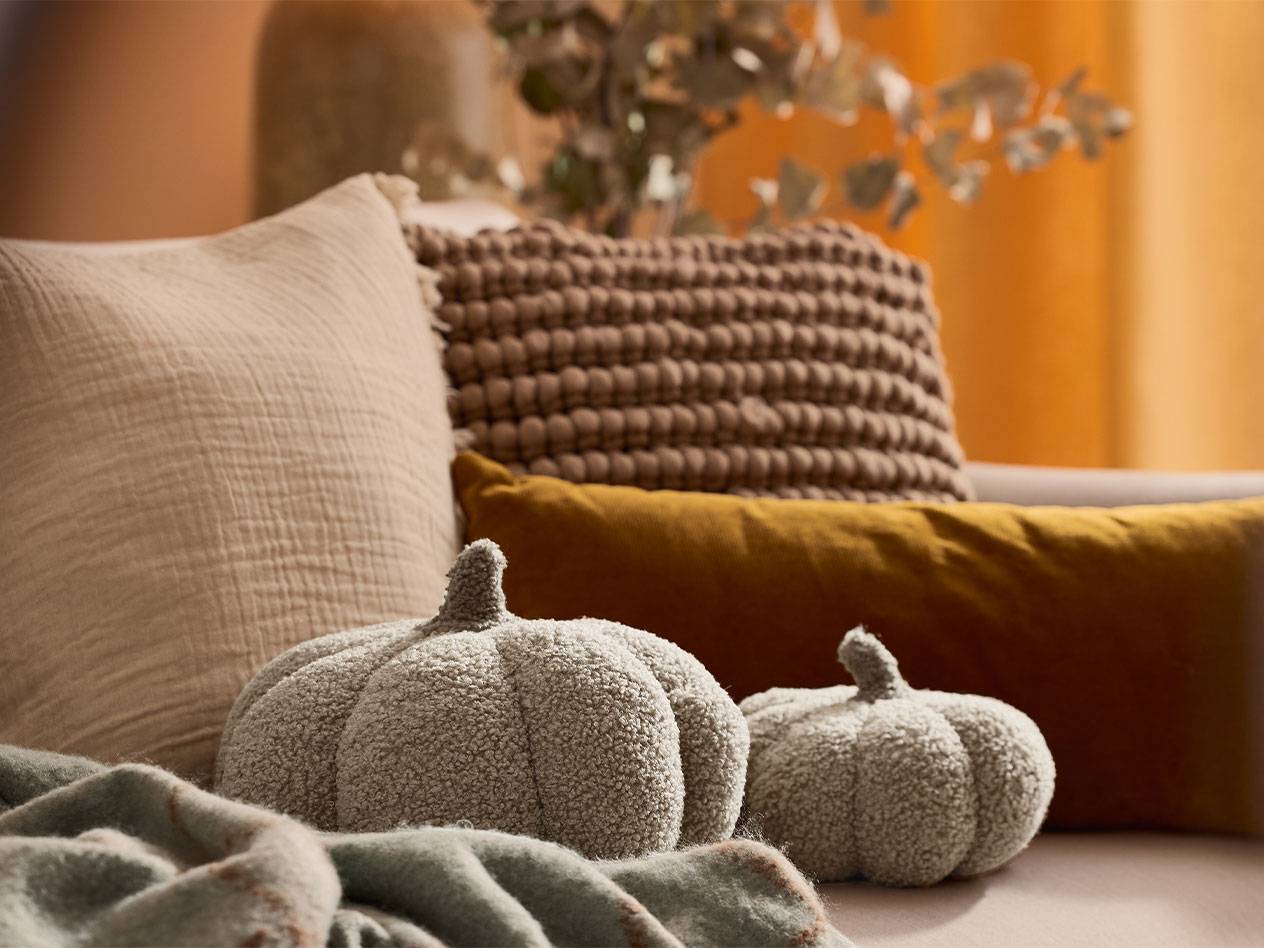 Zwei plüschige Deko-Kürbisse und Kissen in Erdtönen auf einem Sofa.