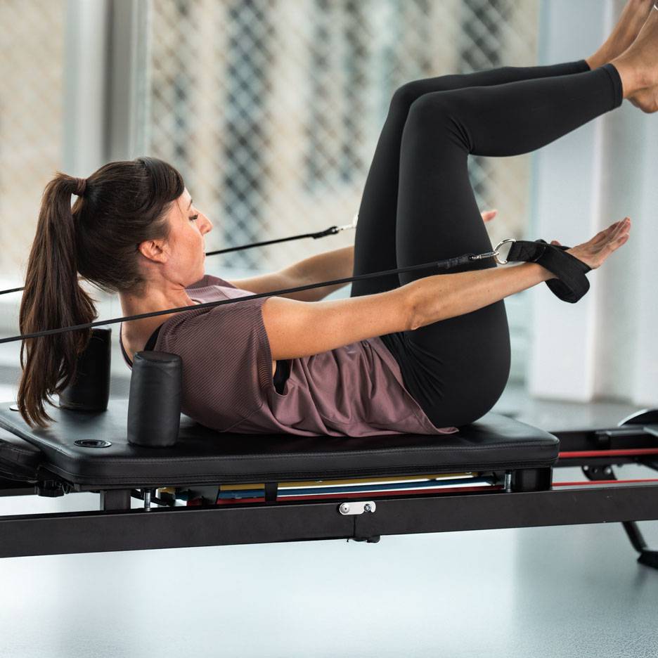 Frau macht Pilates auf einem Reformer, in schwarzer Sportkleidung.