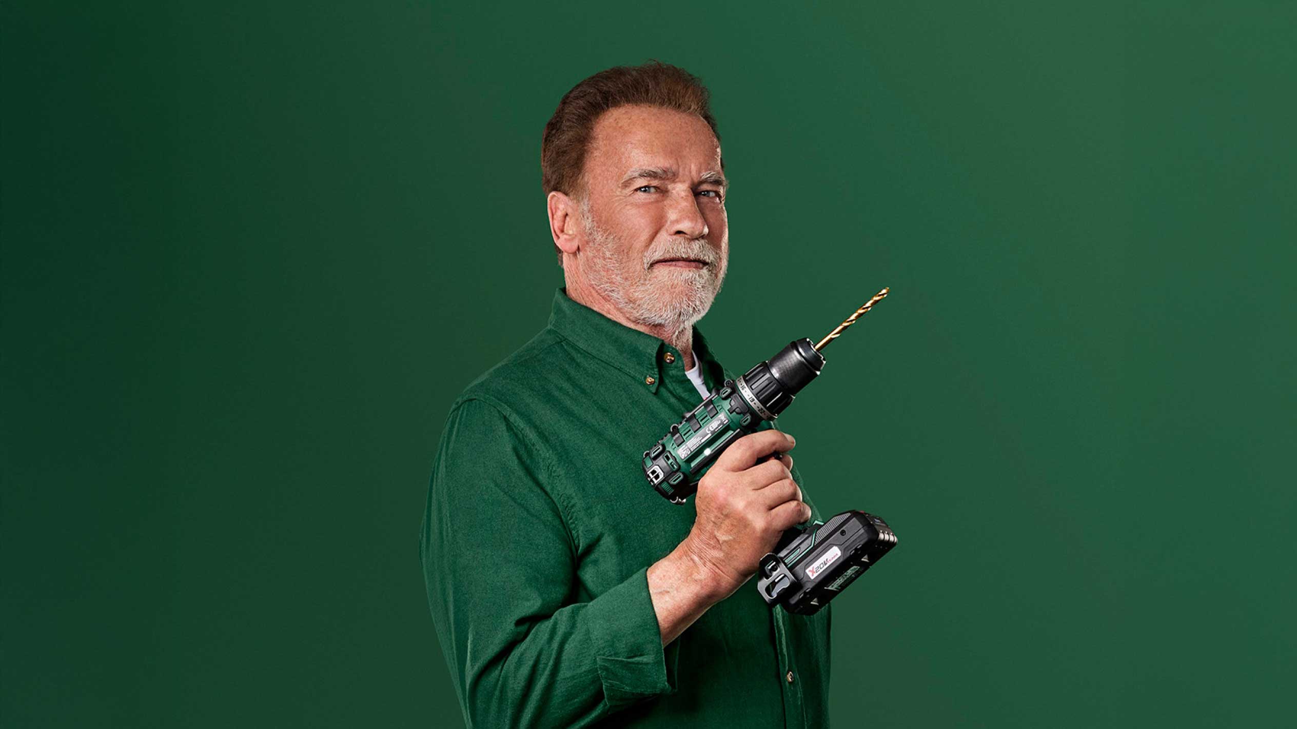 Arnold Schwarzenegger hält einen grünen Akkuschrauber mit goldenem Bohrer.