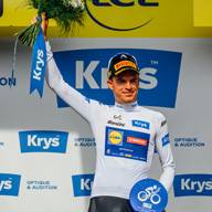 Freudiger Radfahrer in weißem Trikot mit Blumenstrauß und blauem Band auf dem Podium.