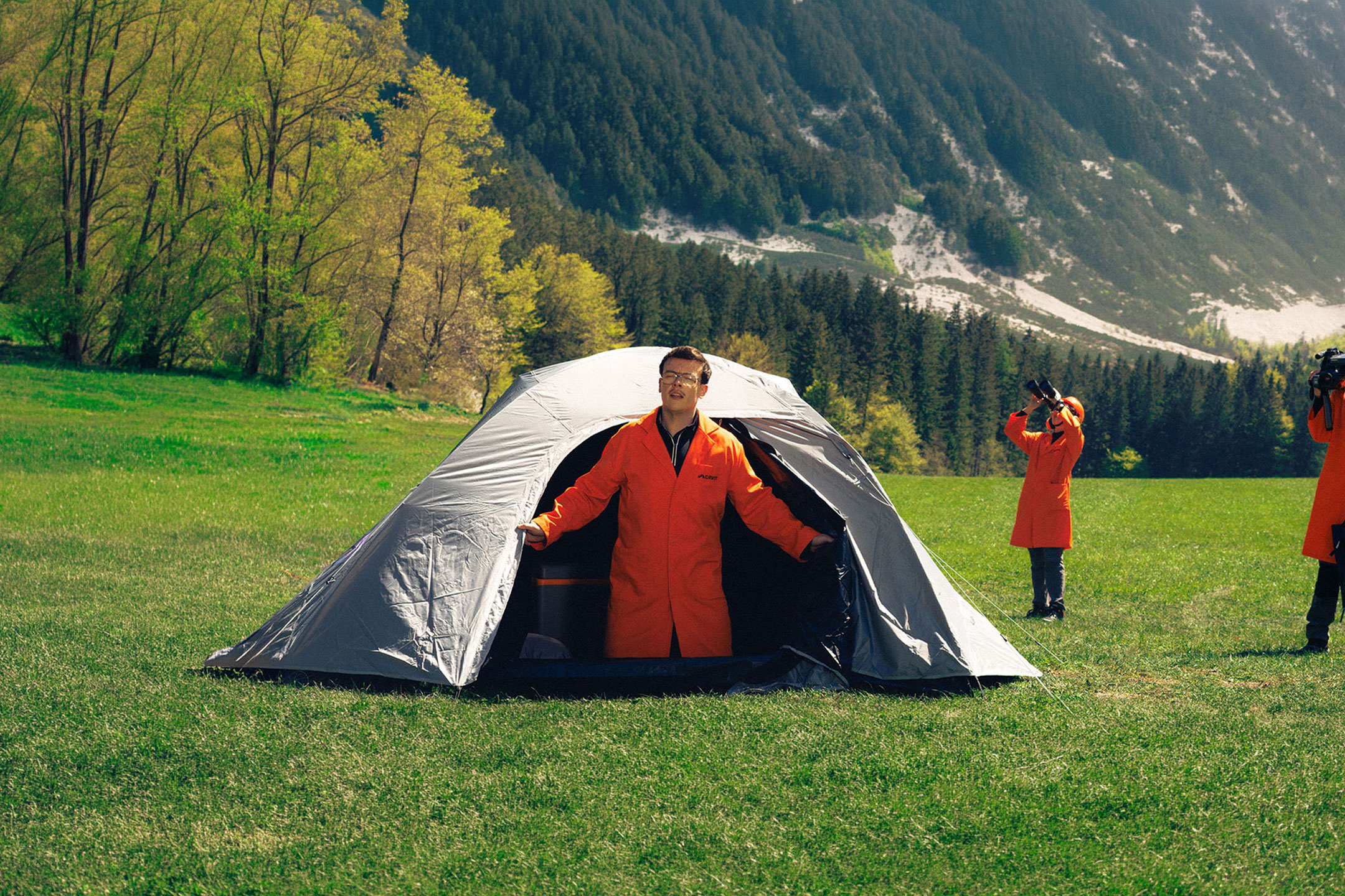 AI-generiert: Drei Personen in orangefarbenen Kitteln mit Camping- und Beobachtungsausrüstung auf einer Wiese.