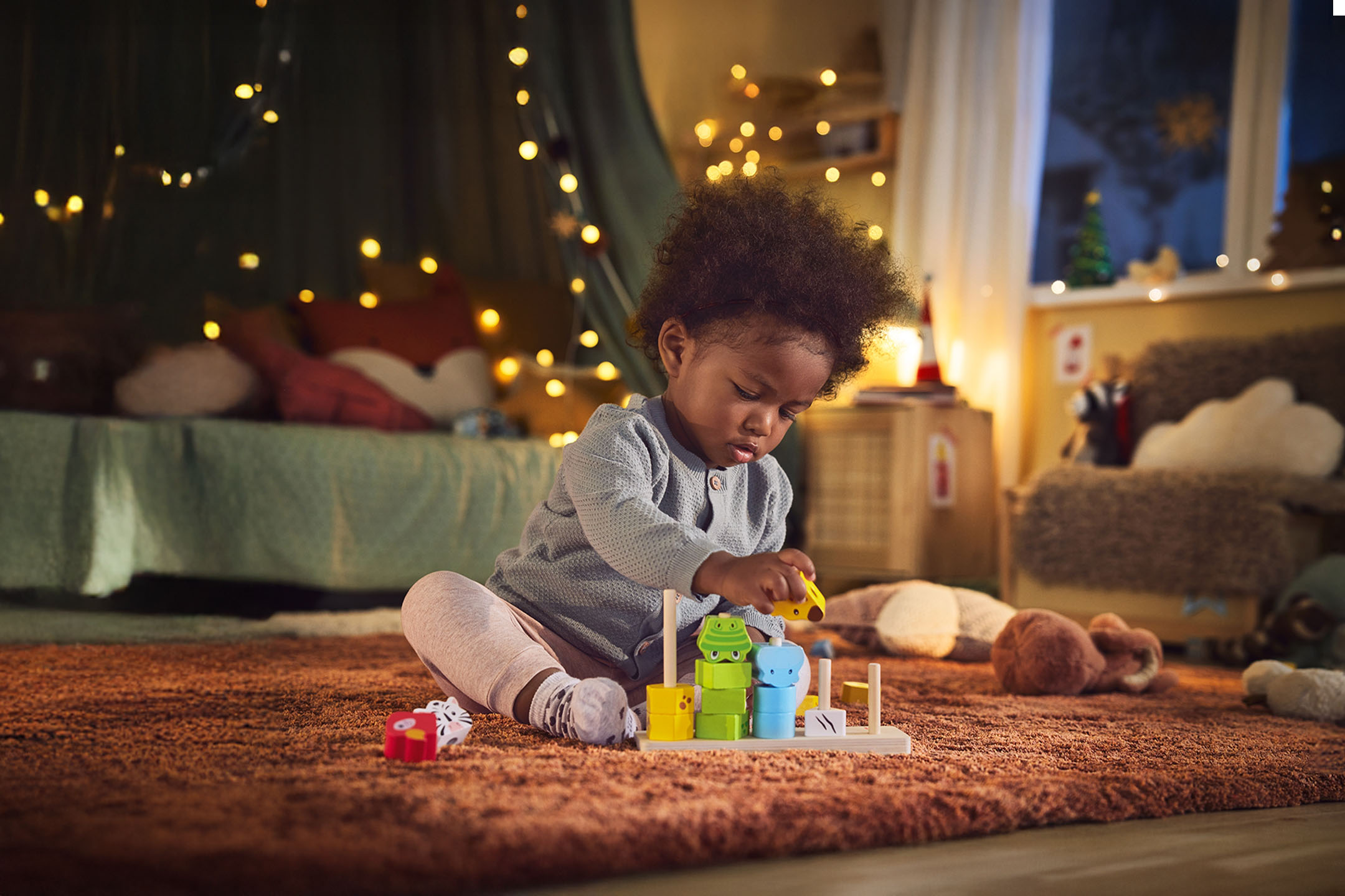 Baby spielt mit Holz-Stapelspielzeug auf flauschigem Teppich, Weihnachtslichter im Hintergrund.