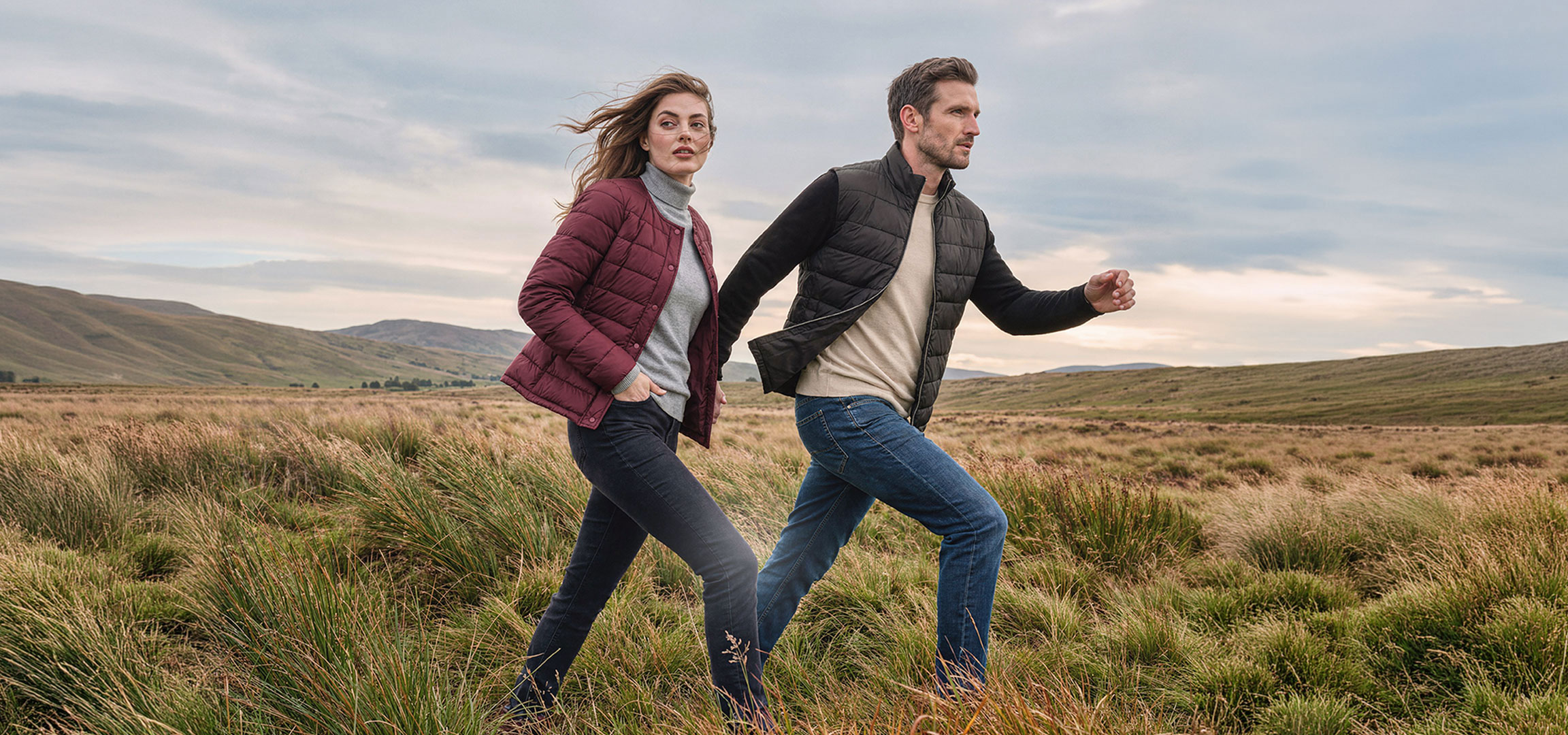 AI-generiert: Paar in leichten Jacken und Jeans geht durch eine grasbewachsene Hügellandschaft.