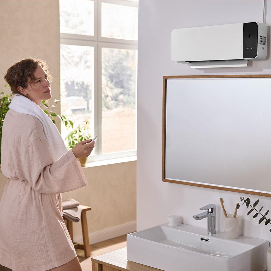 Eine Frau im Bademantel bedient einen Wandheizlüfter im Badezimmer mit einer Fernbedienung.