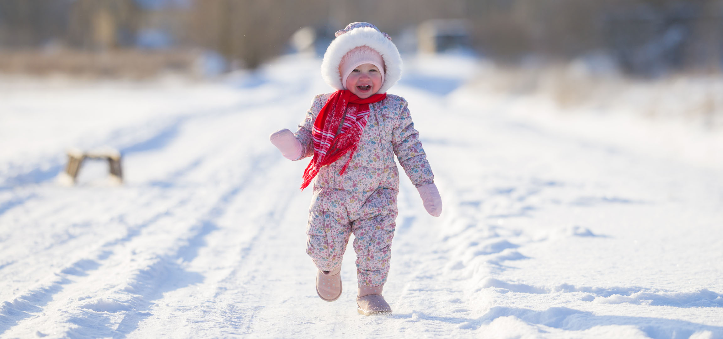 Fröhliches kleines Mädchen in Winteroverall und Schal rennt auf einem verschneiten Weg.