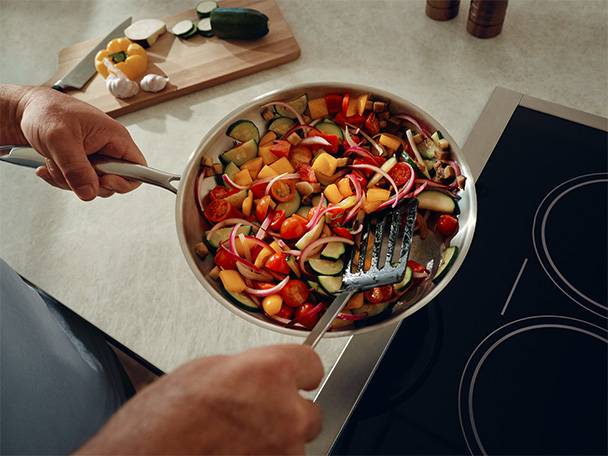 Pfanne mit geschnittenem Gemüse wie Zucchini, Paprika, Zwiebeln und Tomaten auf einem Induktionskochfeld.
