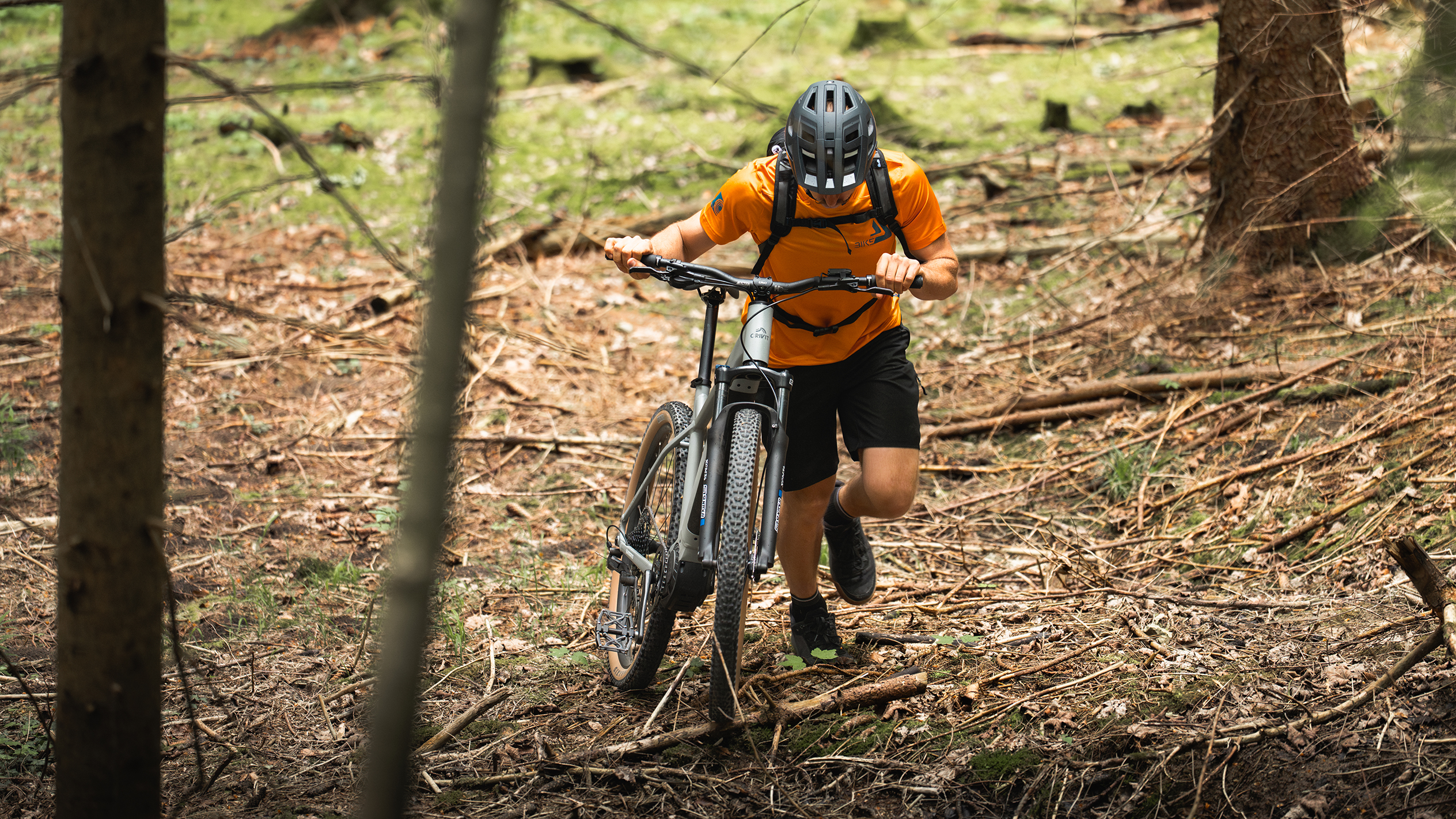 Ein Radfahrer in orangem Shirt schiebt ein E-Mountainbike durch einen Wald.