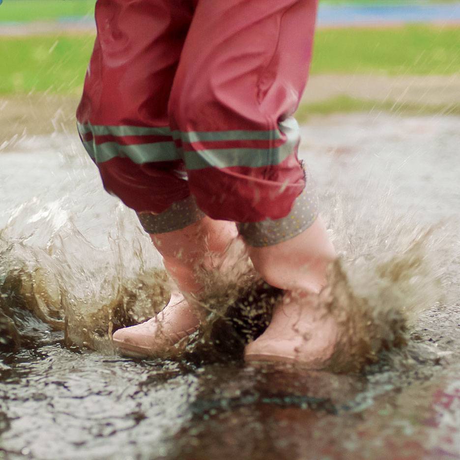 Kind in roter wasserdichter Hose und Stiefeln springt in eine Pfütze.
