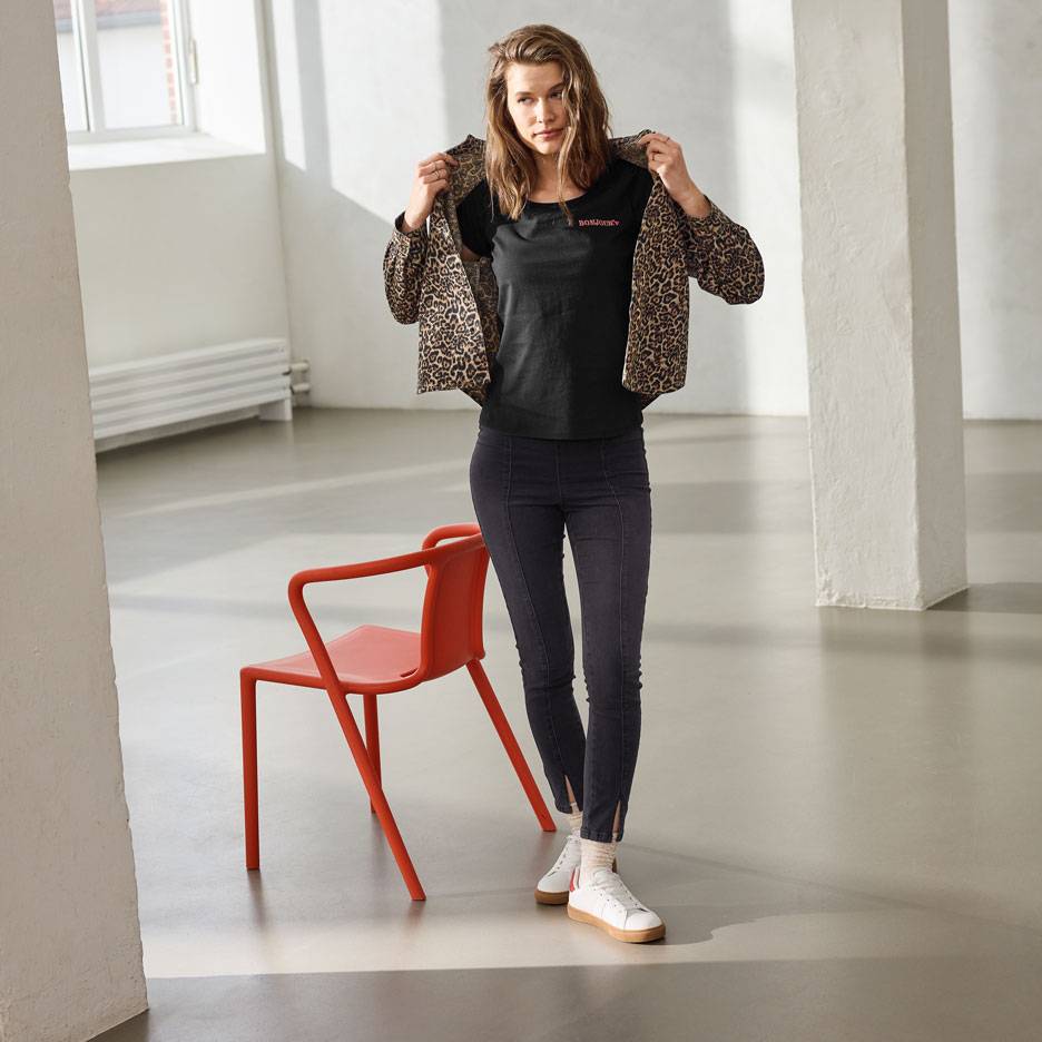 Frau in schwarzem T-Shirt mit Text, Jeans und Leopardenjacke, steht neben einem roten Stuhl.