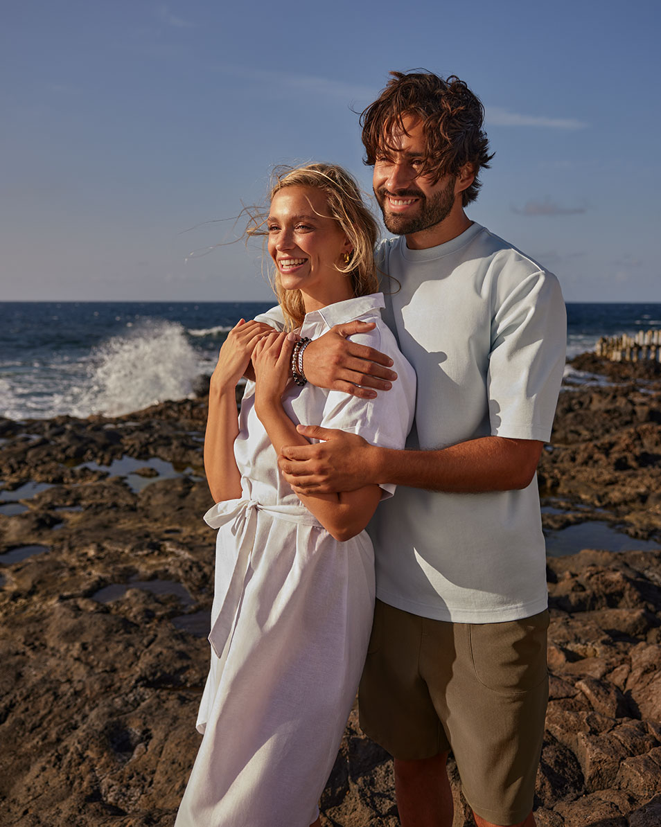 Paar am Strand, Frau in weißem Kleid, Mann in hellblauem T-Shirt und Shorts.