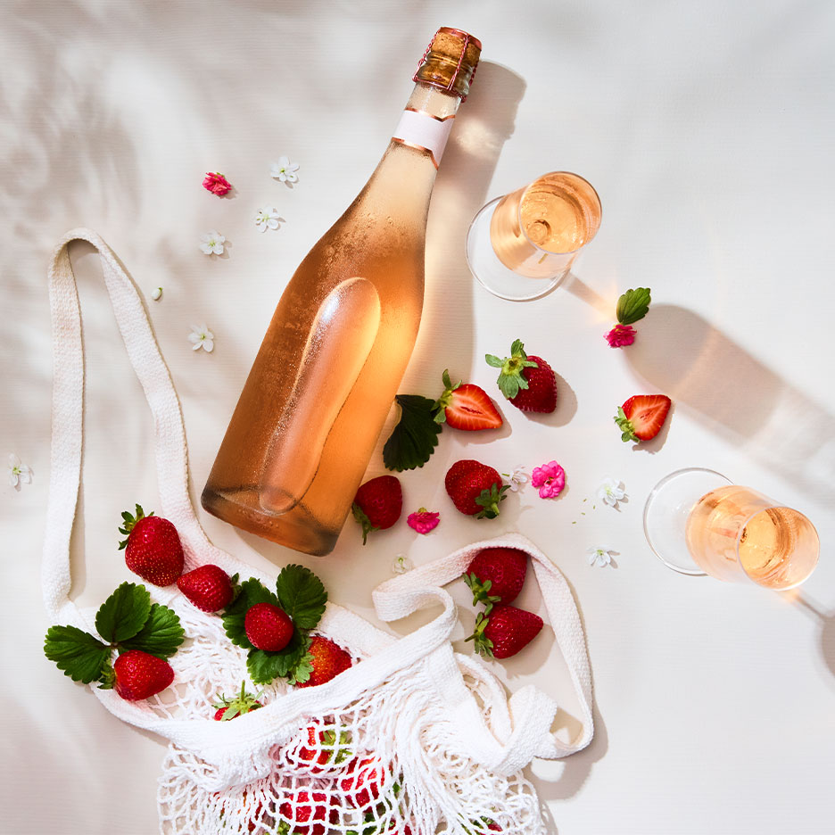 Roséweinflasche und Gläser mit frischen Erdbeeren und Blumen auf einer weißen Oberfläche.