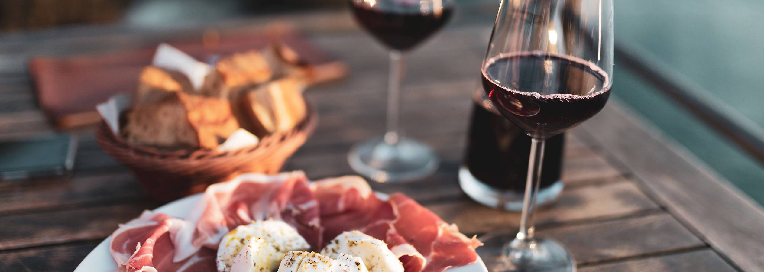 Ein Teller mit Schinken und Mozzarella, dazu Brot und zwei Gläser Rotwein auf einem Holztisch.