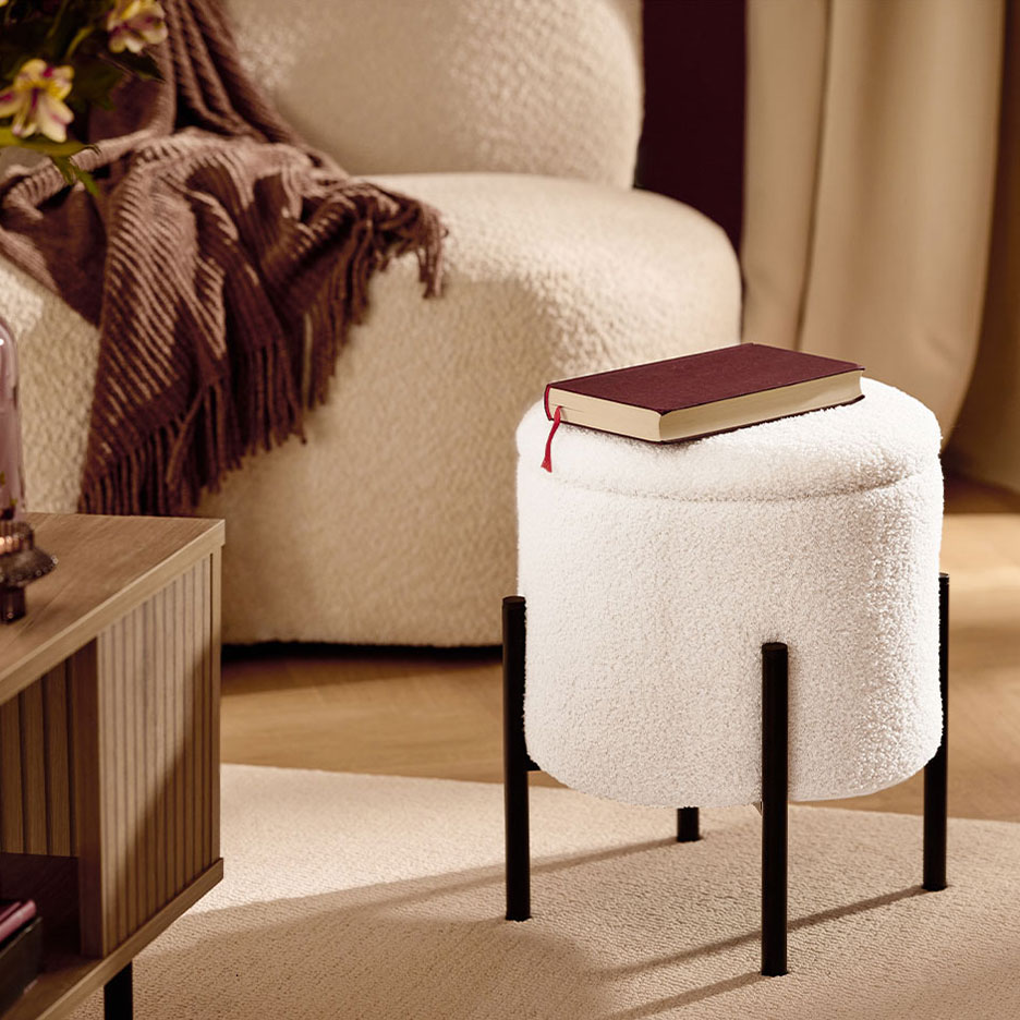 Weißer flauschiger Hocker mit schwarzen Beinen, ein rotes Buch, Sessel und braune Decke.