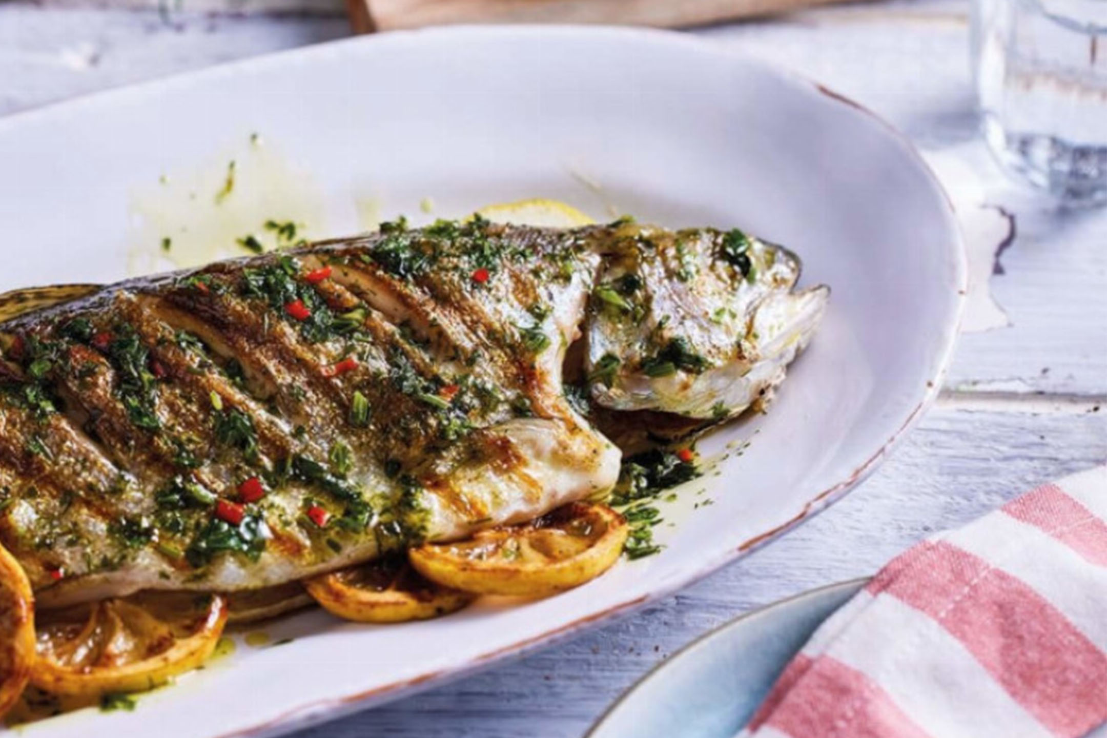 Gegrillter ganzer Fisch mit Kräutern und Zitrone auf einer weißen Platte.