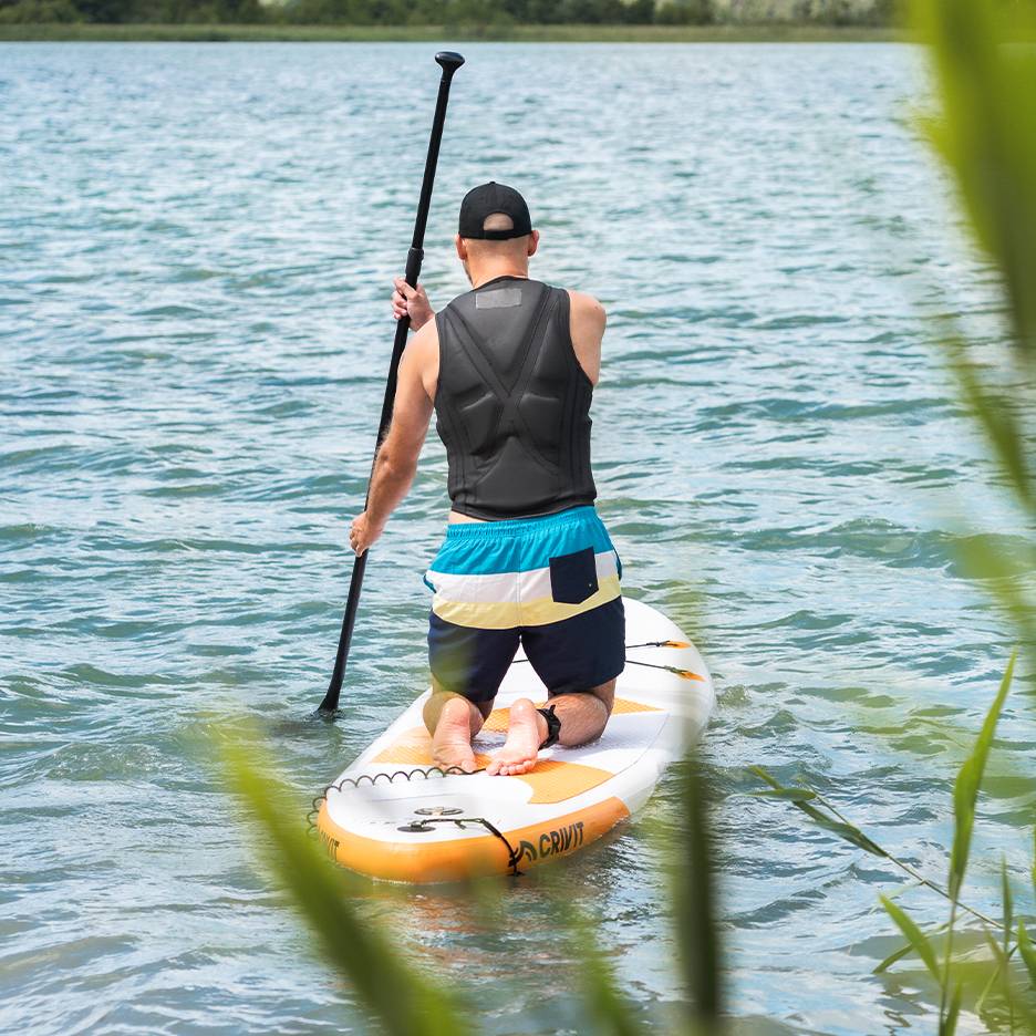 Mann paddelt auf einem See auf einem Crivit SUP-Board, mit Schwimmweste und Badeshorts.