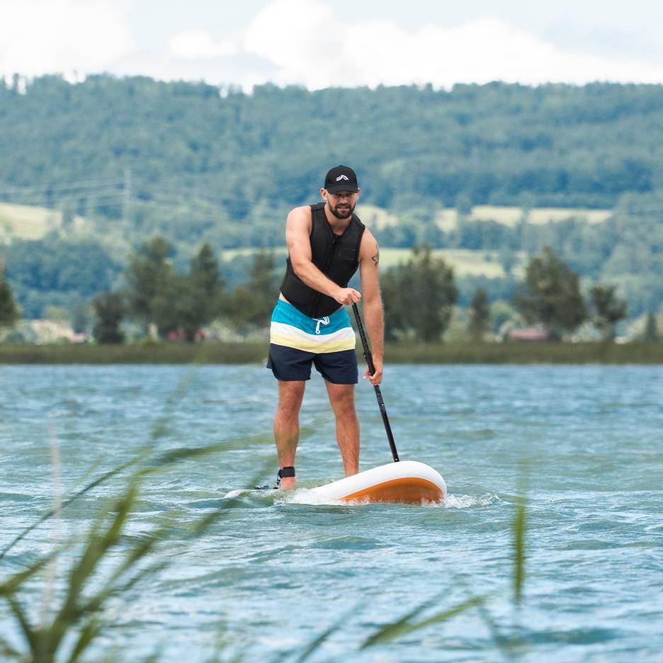 Mann beim Stand-Up-Paddling auf einem See, mit Schwimmweste und Badeshorts.
