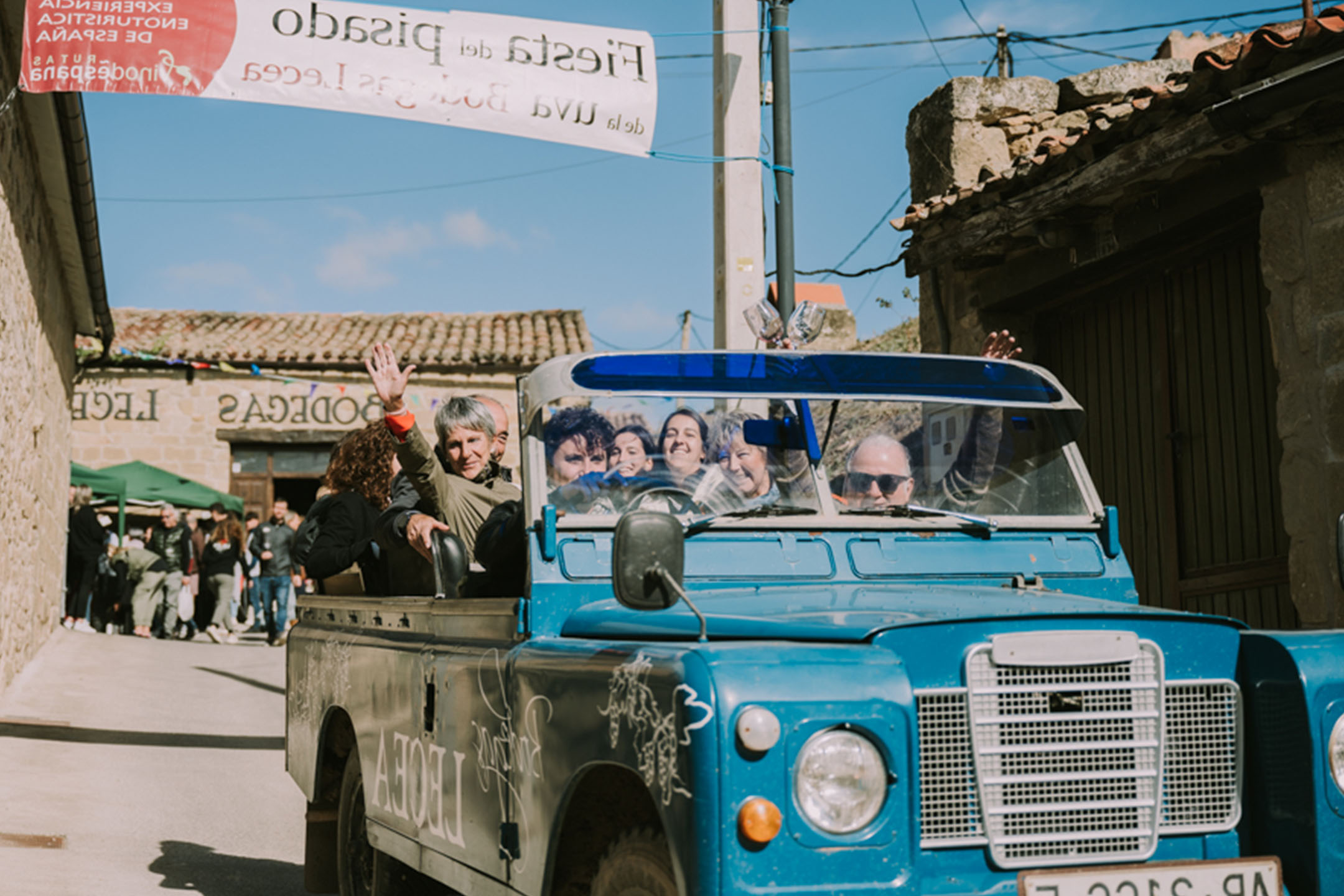 Menschen in einem blauen Geländewagen winken auf einer Straße mit einem „Fiesta del Pisado“-Banner.