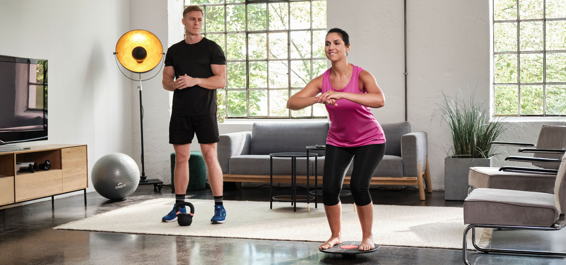Frau und Mann trainieren zu Hause mit Balance Board und Kettlebell.