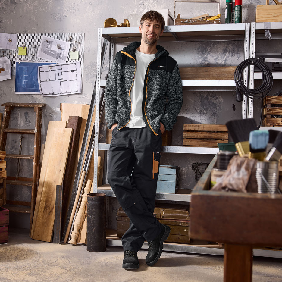 Mann in Fleecejacke und Arbeitshose in einer Werkstatt.