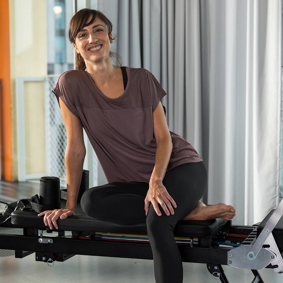 Frau in Sportkleidung lächelt, sitzend auf einem Pilates-Gerät.