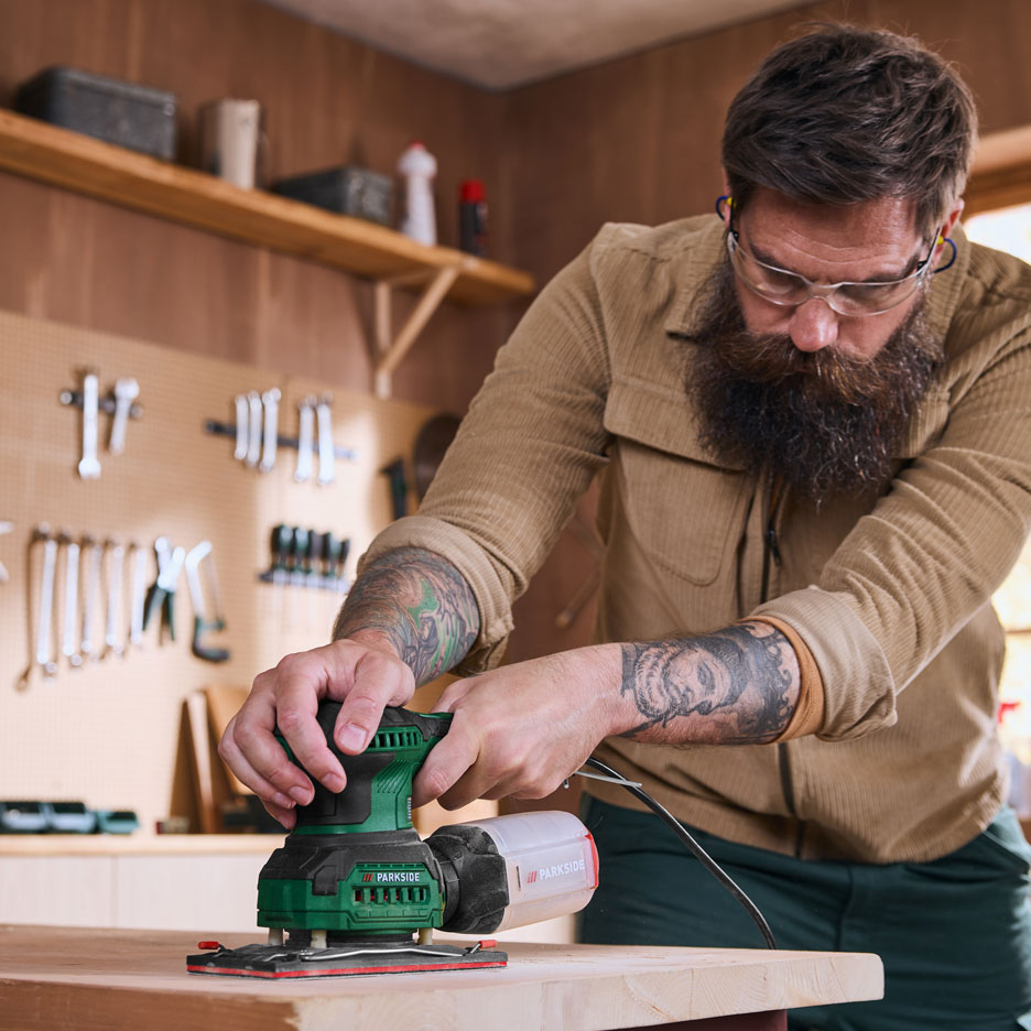 Mann in Werkstatt schleift ein Holzbrett mit einem Elektroschleifer.