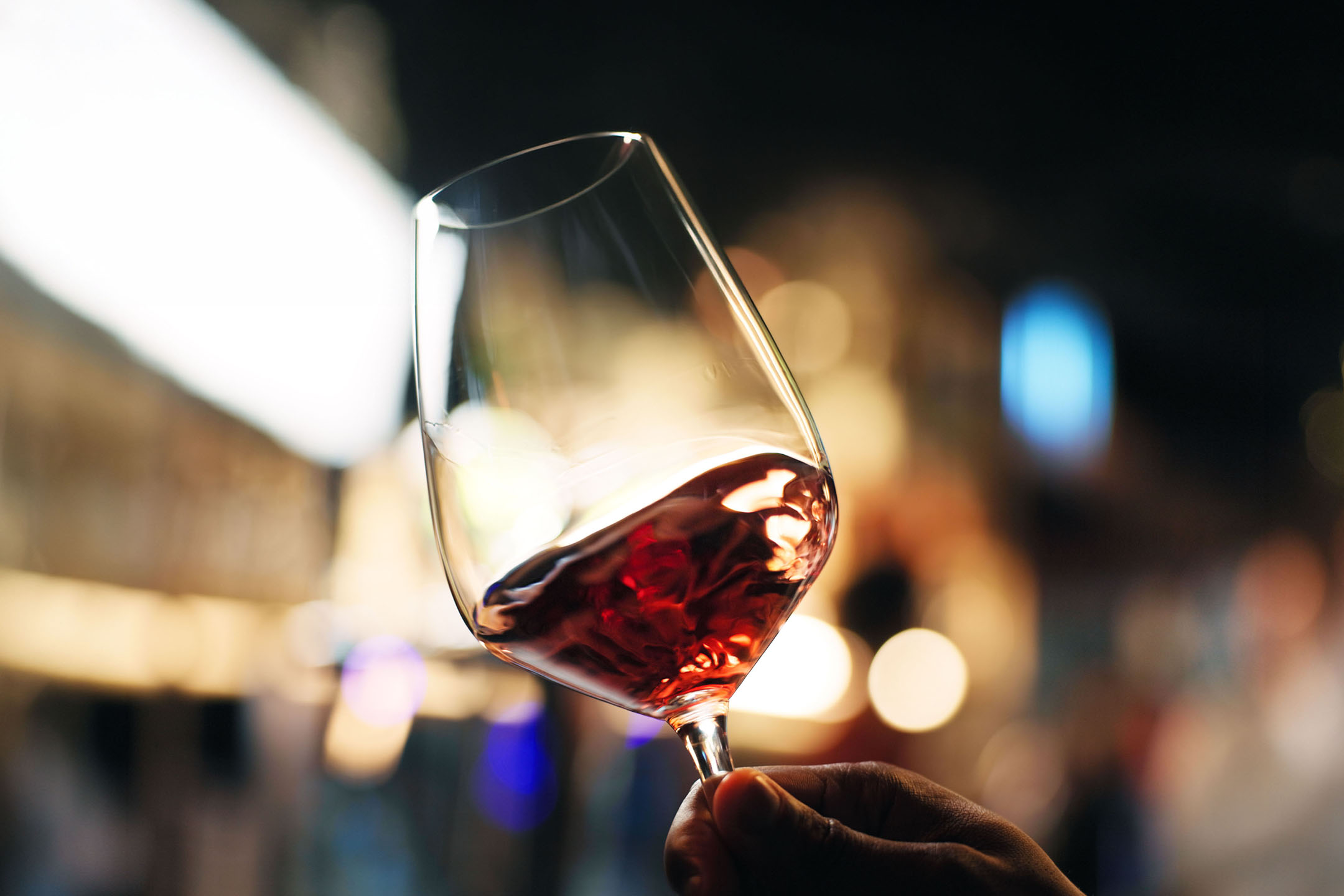 Glas Rotwein in der Hand, mit verschwommenen Lichtern im Hintergrund.