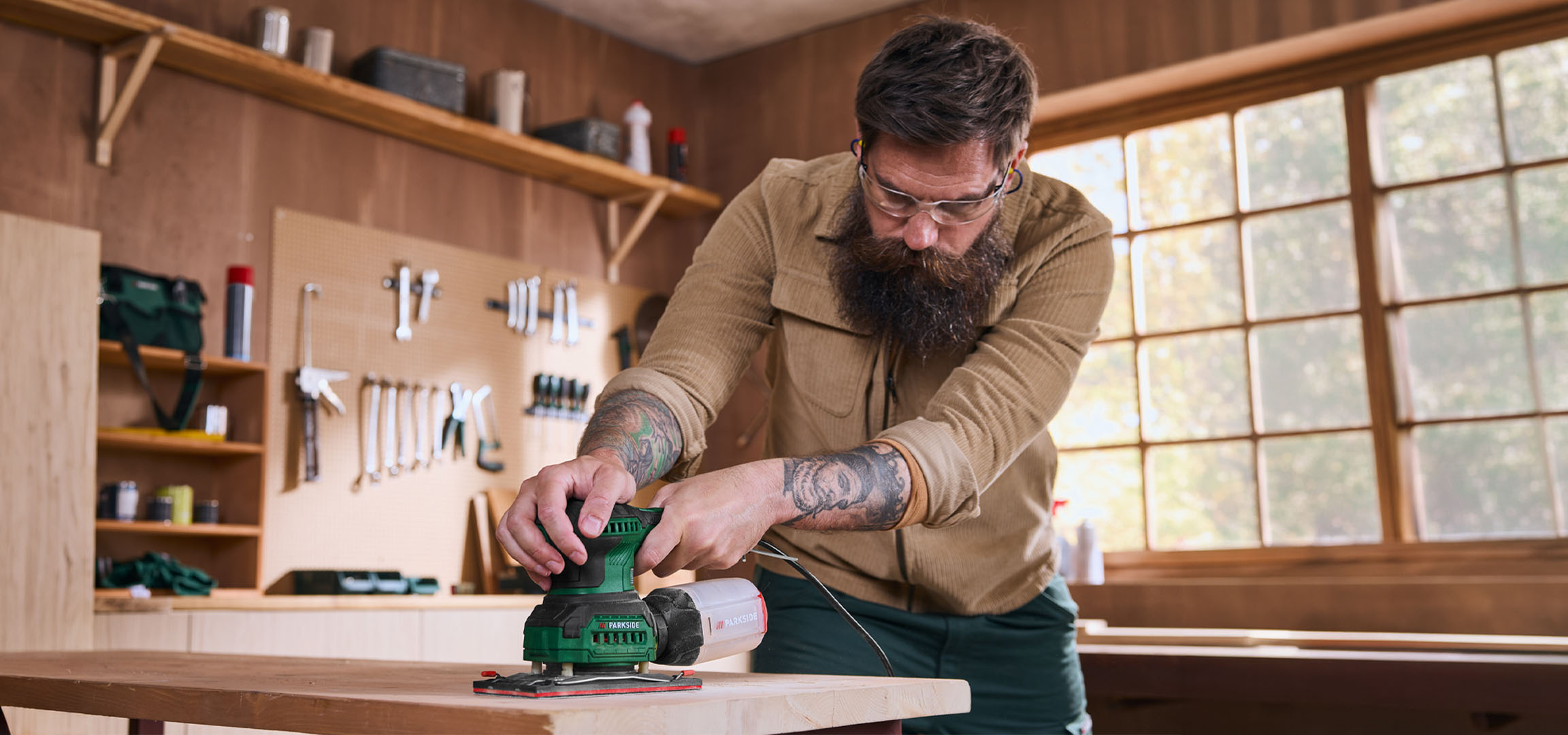 Mann schleift ein Holzbrett mit einem Elektroschleifer in einer Werkstatt.