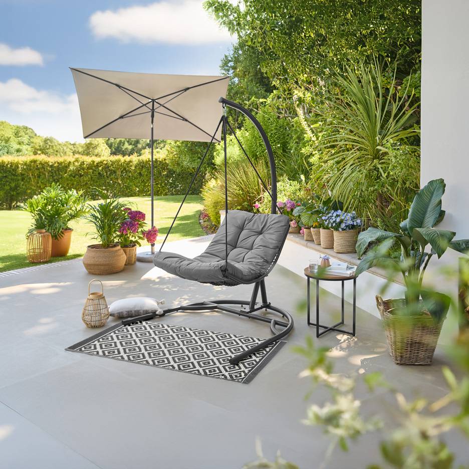 Grauer Hängesessel mit Kissen, gemusterter Teppich und Sonnenschirm auf einer grünen Gartenterrasse.