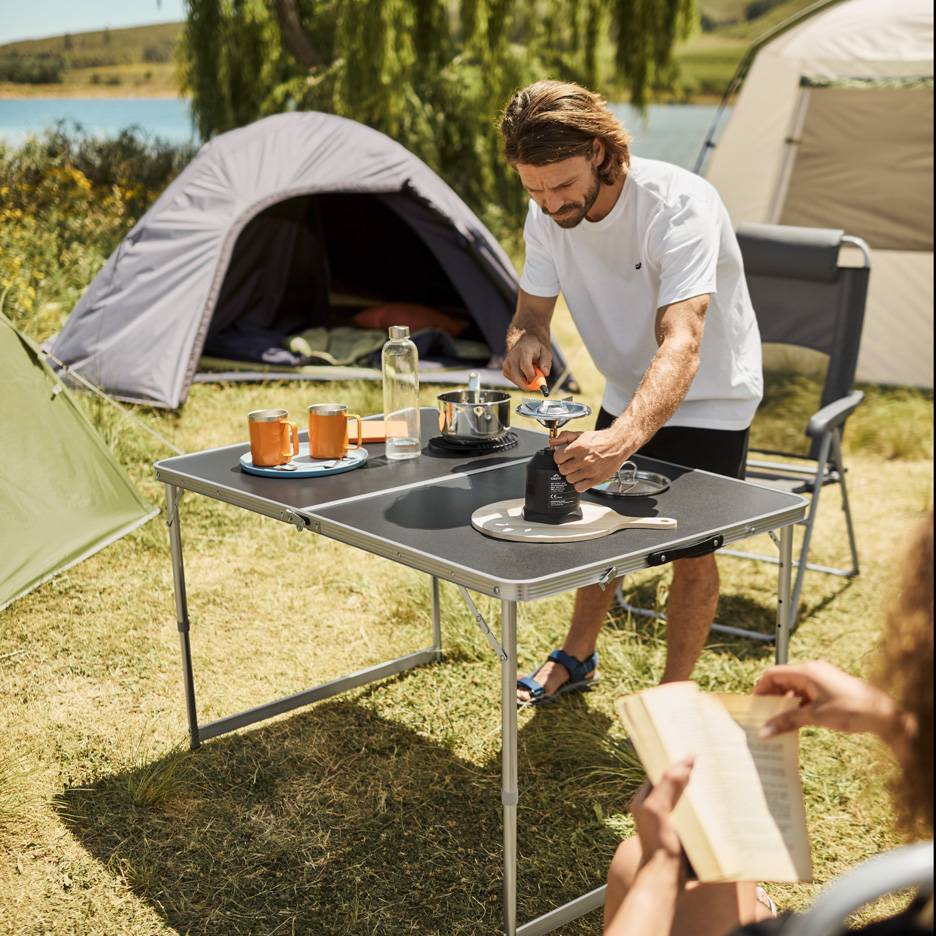 Mann bereitet Campingmahlzeit auf Klapptisch mit Kocher und Tassen zu.