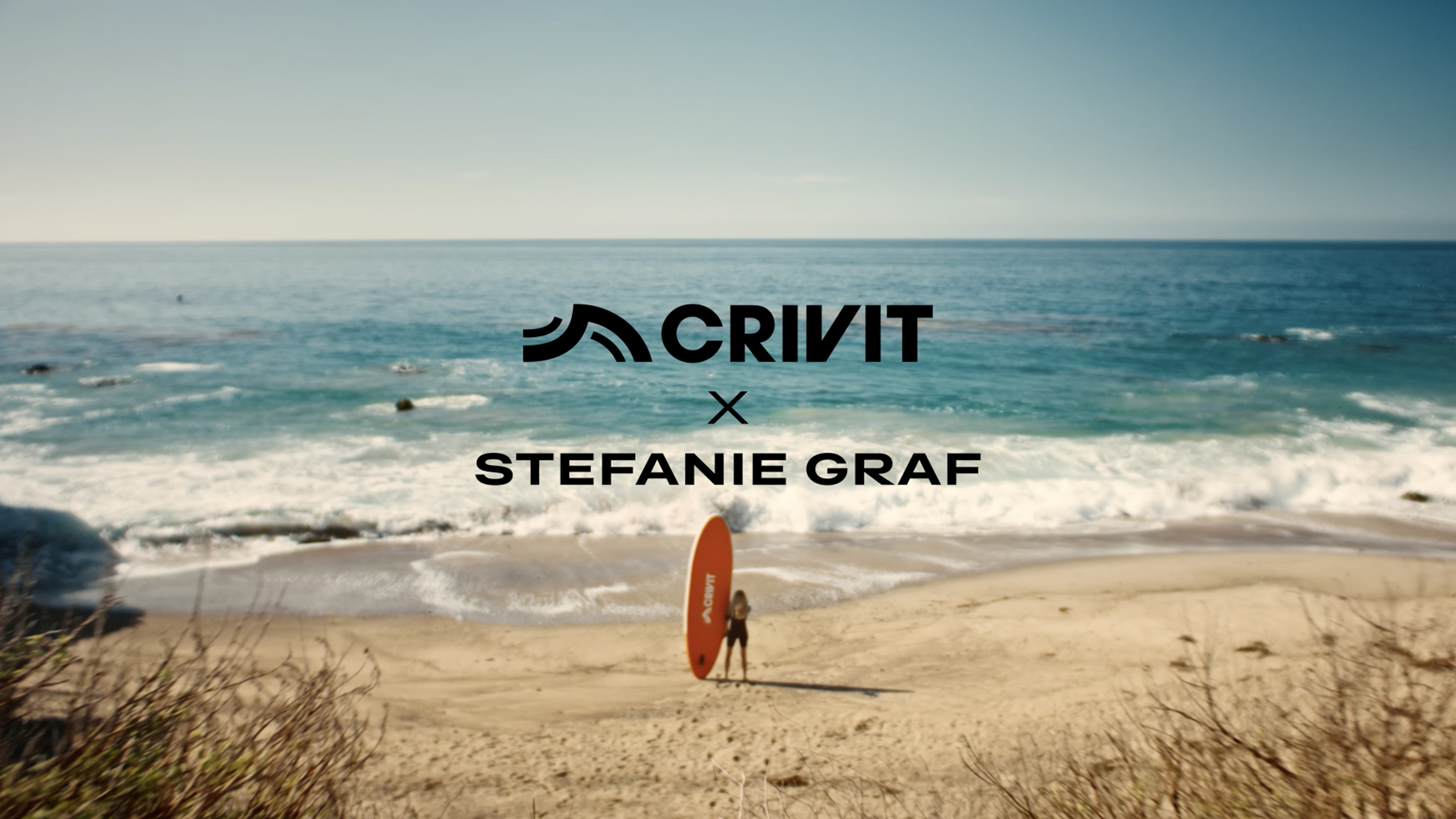 Frau mit orangefarbenem SUP-Board an einem Sandstrand, mit dem Meer im Hintergrund.