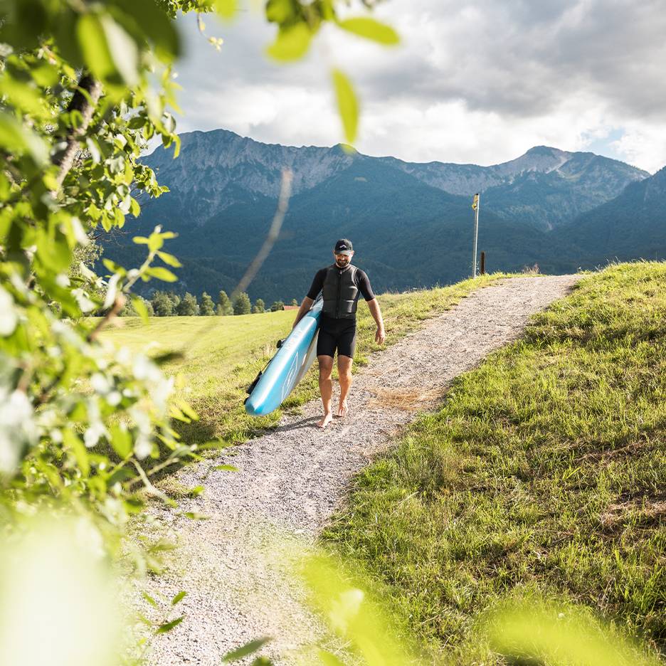 Mann in Neoprenanzug und Schwimmweste trägt ein Paddleboard in Berglandschaft.