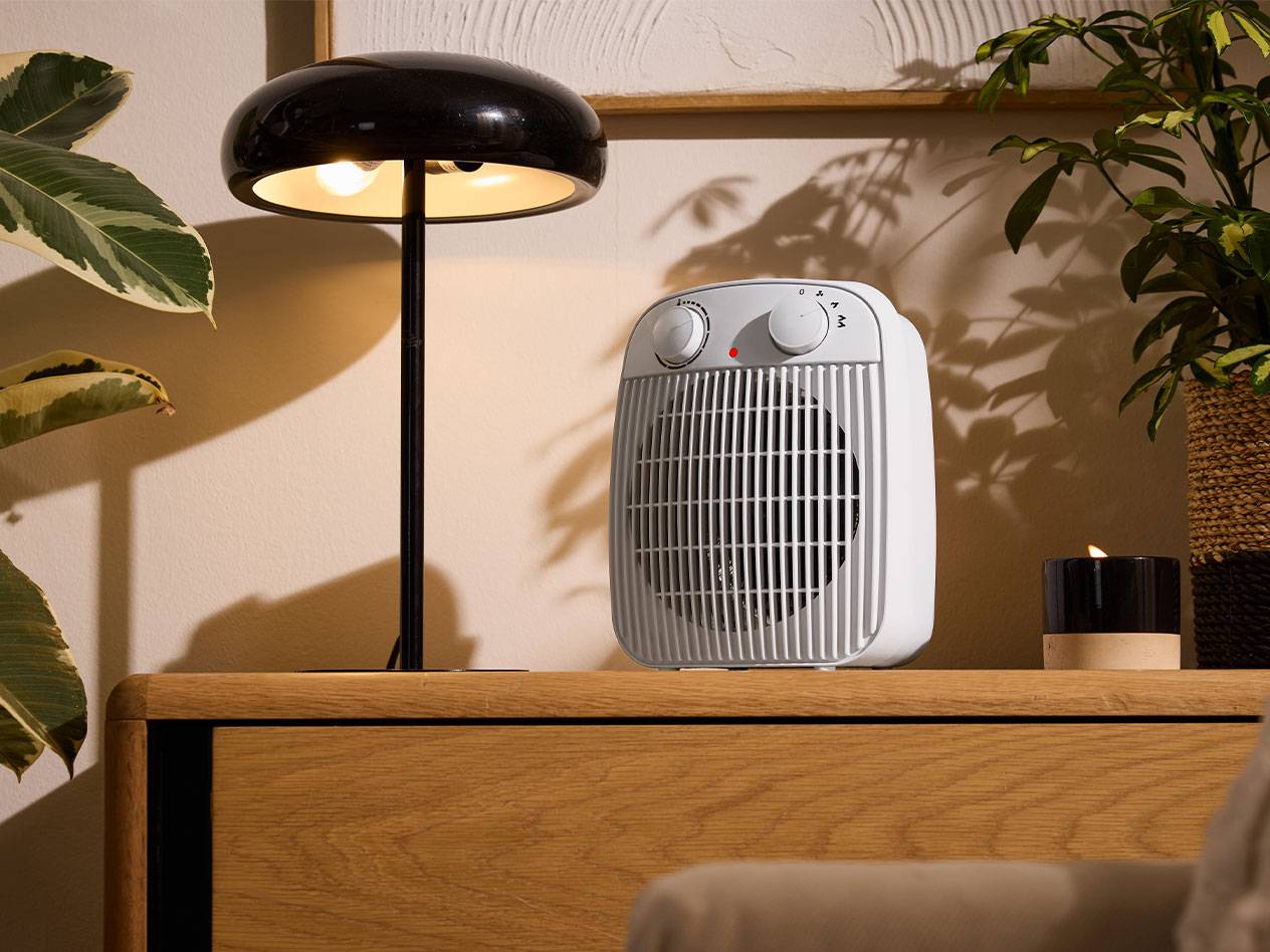 Weißer Heizlüfter und schwarze Tischlampe auf einem Holzmöbel in einem gemütlichen Raum.
