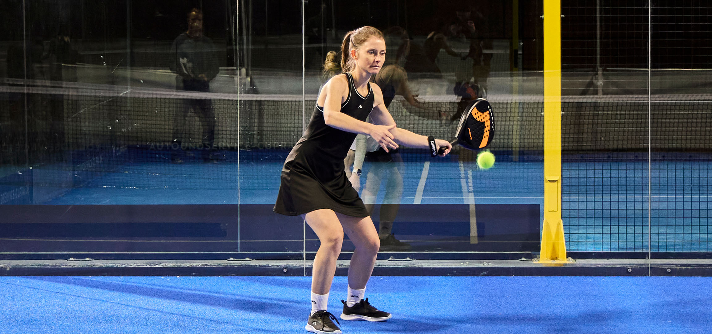 Frau spielt Padel auf blauem Platz, gekleidet in schwarzer Sportkleidung.