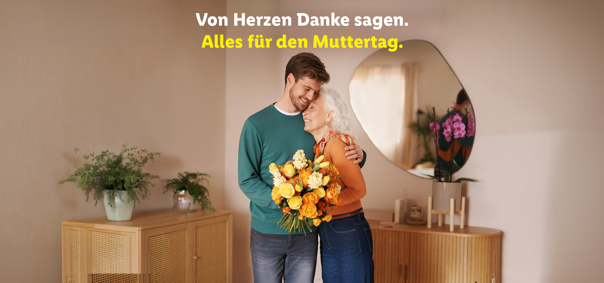 Ein junger Mann umarmt eine ältere Frau mit einem Blumenstrauß, mit dem Text „Von Herzen Danke sagen. Alles für den Muttertag.“