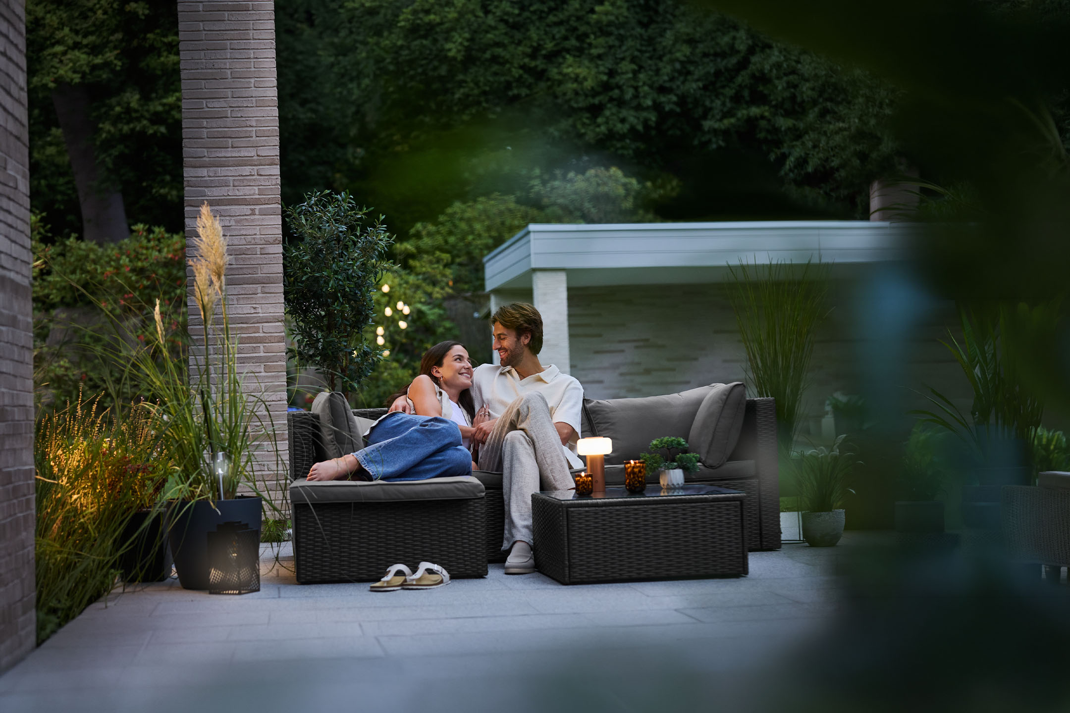 Paar entspannt auf einem Outdoor-Sofa auf einer Terrasse mit Gartenmöbeln und Beleuchtung.