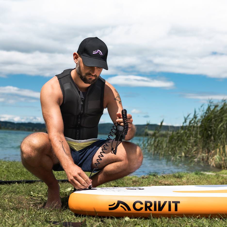 Mann befestigt Leash am CRIVIT SUP-Board, trägt Schwimmweste und Kappe.
