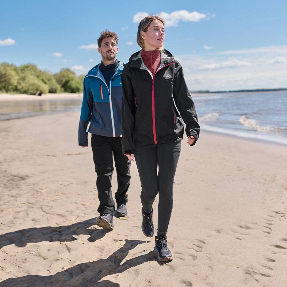 Paar in Wanderjacken und -hosen geht an einem Sandstrand am Meer spazieren.