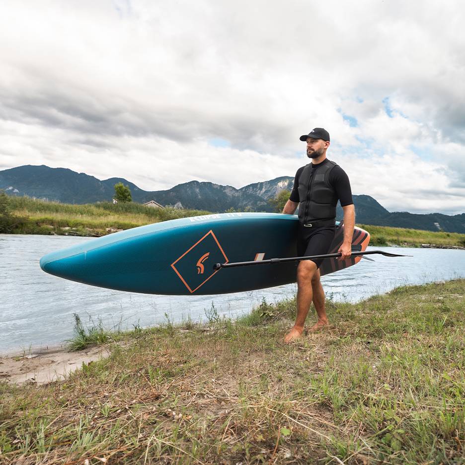 Mann in Schwimmweste und Kappe trägt ein Paddleboard und Paddel am Fluss.