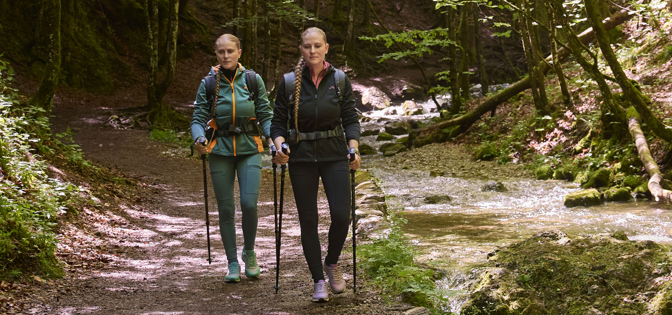 Zwei Frauen wandern mit Wanderstöcken entlang eines Baches in einem Wald.